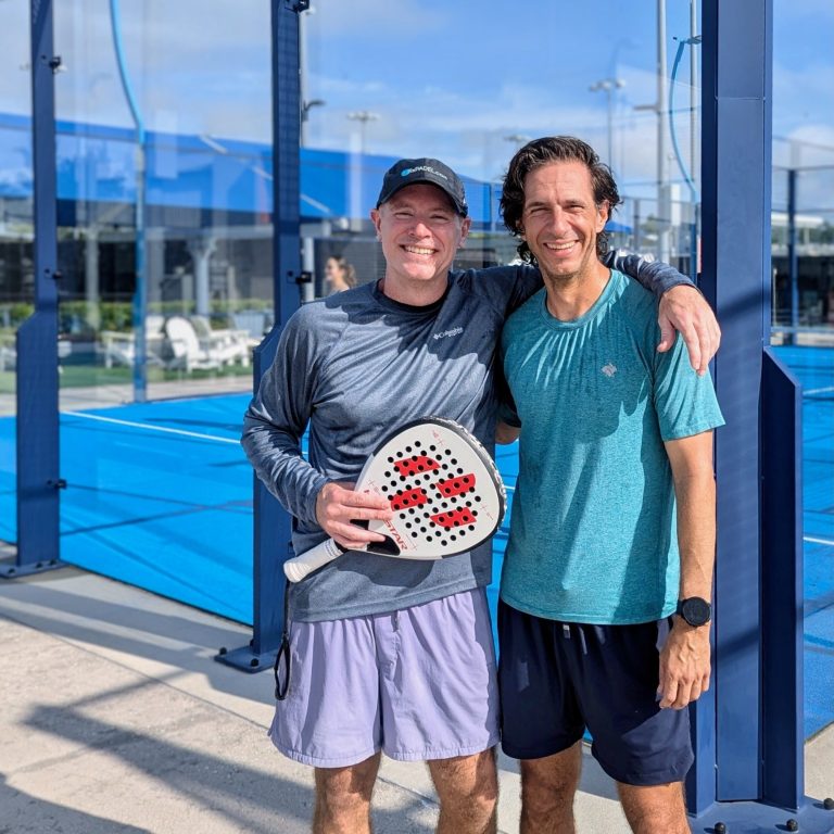 All Levels Welcome Two padel players, one holding pala or padel racquet, pause outside the court for a photo together.